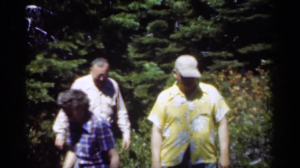 1949 New Orleans home-movie frame showing three adults moving through a wooded setting, including a man in a yellow floral shirt.