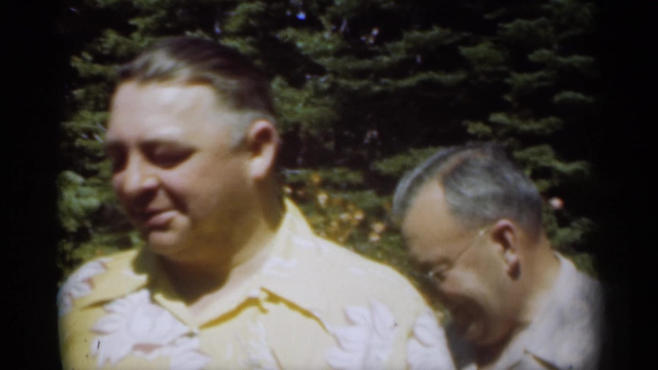 Two adult men stand close together outdoors, one in a yellow shirt and one partly behind him.