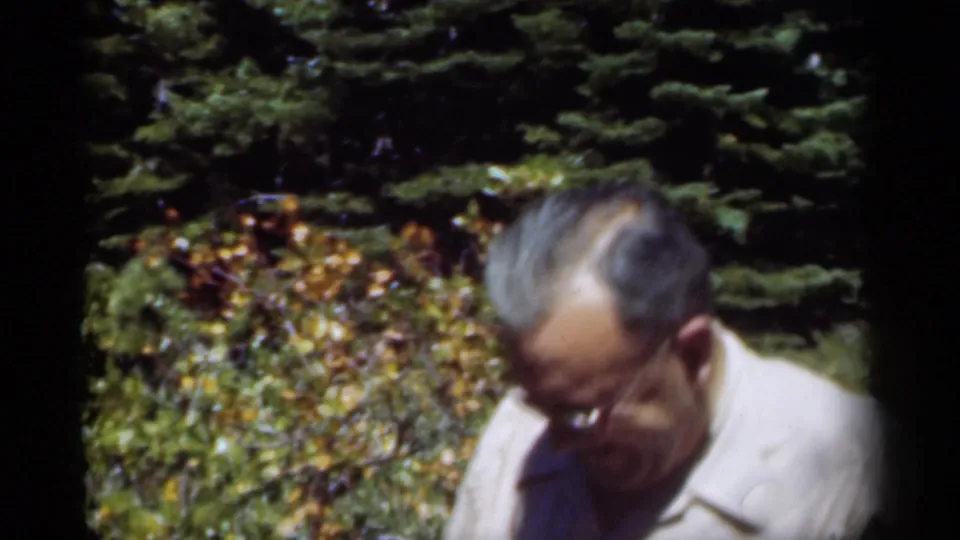 A bespectacled man in a light collared shirt looks down in a leafy New Orleans garden setting.