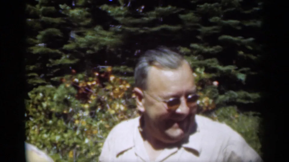 The man in glasses smiles slightly while standing in a shaded, leafy garden in 1949 home-movie footage.