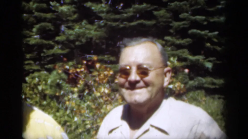 A smiling man in a light collared shirt and glasses faces the camera in a sunlit New Orleans garden scene.