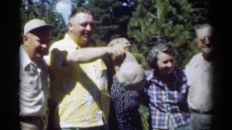 In the 1949 home-movie scene, adults gather in sunlit woods while a man in a yellow patterned shirt playfully holds a light-colored hat.