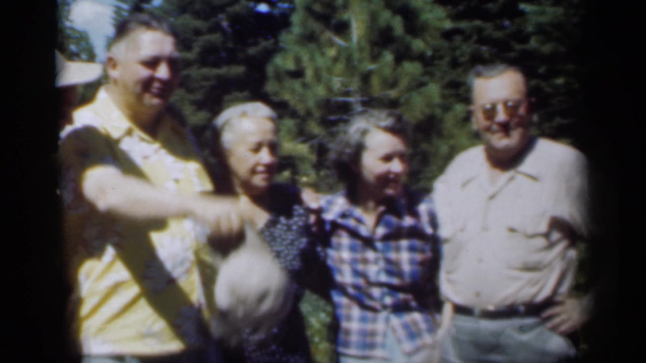 Four adults stand outdoors among tall trees, with a man in a yellow shirt raising his arm.