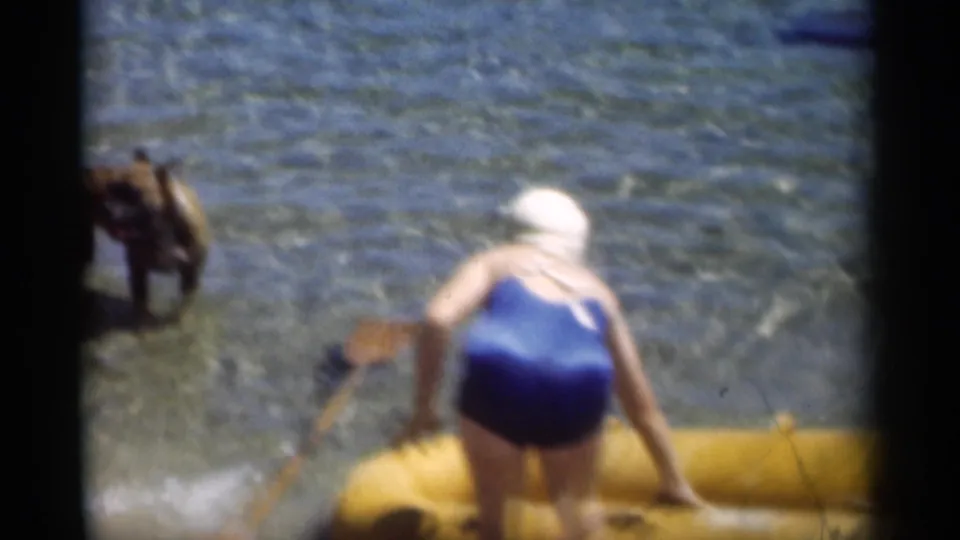 1949 New Orleans-area home-movie frame showing a woman in a blue swimsuit stepping from a yellow inflatable boat near a dog in shallow water.