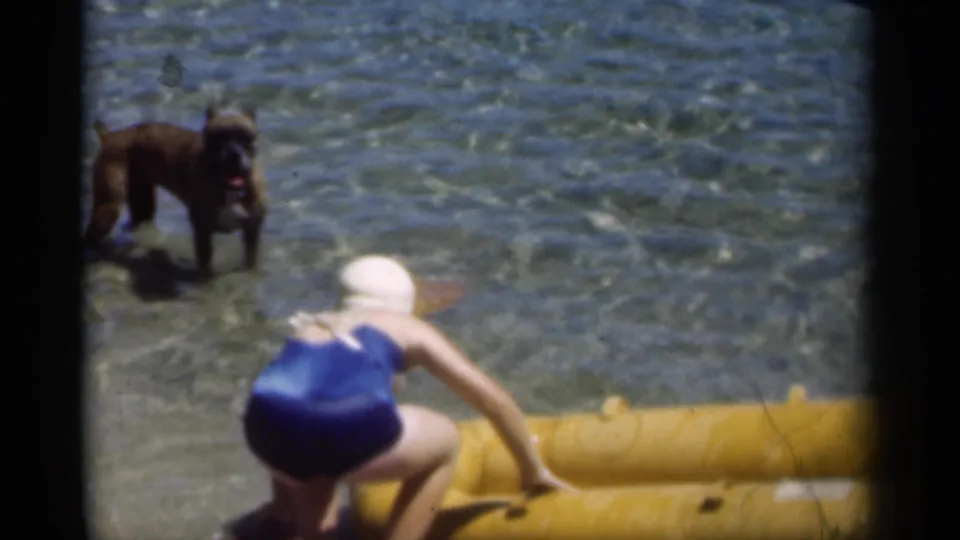 1949 New Orleans-area beach frame with a woman in swimwear crouched on a yellow inflatable boat and a dog standing in the surf.