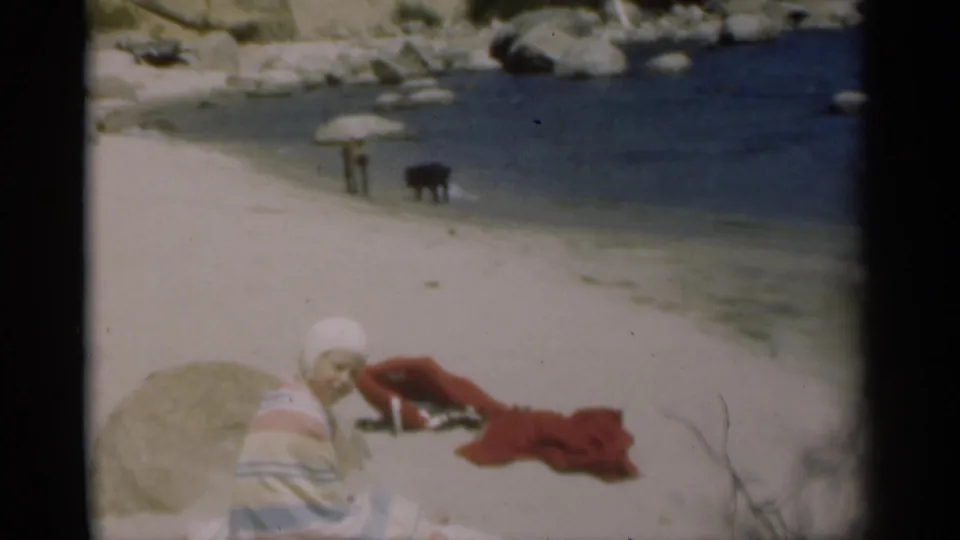 The seated girl turns her face toward the camera as two dogs stand near the shoreline in the 1949 home-movie frame.