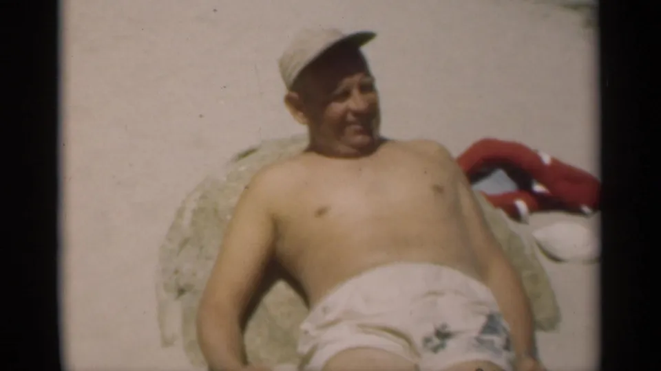 A shirtless adult man in a cap reclines on the sandy beach, with towels or clothing nearby, in 1949 home-movie footage.
