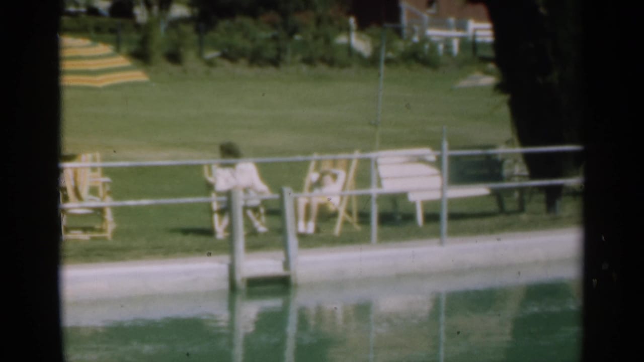 Two seated figures beside an outdoor swimming pool with lawn, chairs, and railings.