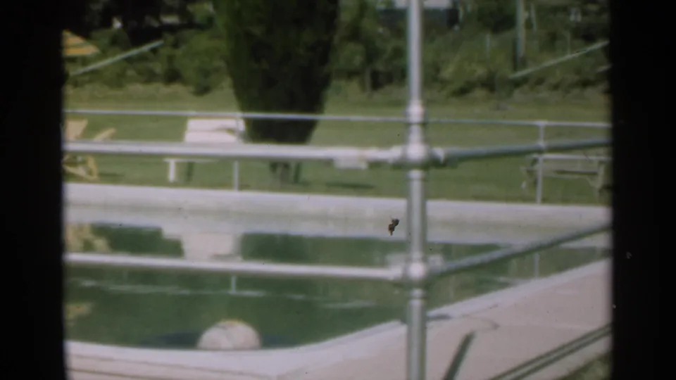 Santa Cruz 1949 home-movie frame focused on the pool edge, metal railings, reflected water, lawn, deck chairs, and trees.