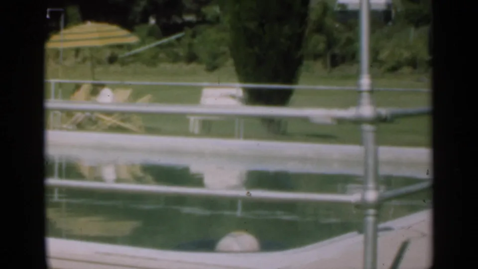 Santa Cruz 1949 poolside frame showing water, railings, lawn, wooden chairs, a striped umbrella, and trees in soft 8mm color.