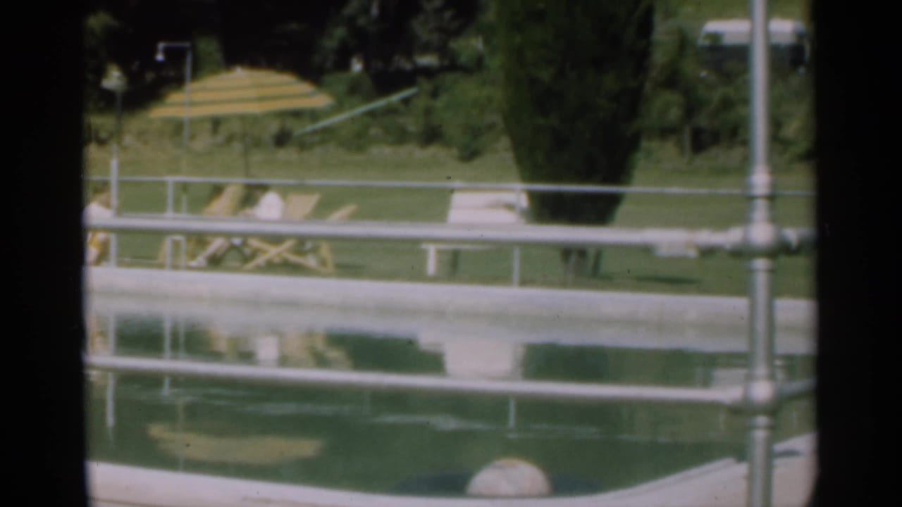 Poolside view with railing, lawn, striped umbrella, lounge chair, and two people near the water.