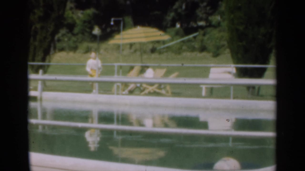 1949 Santa Cruz poolside frame with a standing figure near the water, a striped umbrella, lounge chair, lawn, and trees.