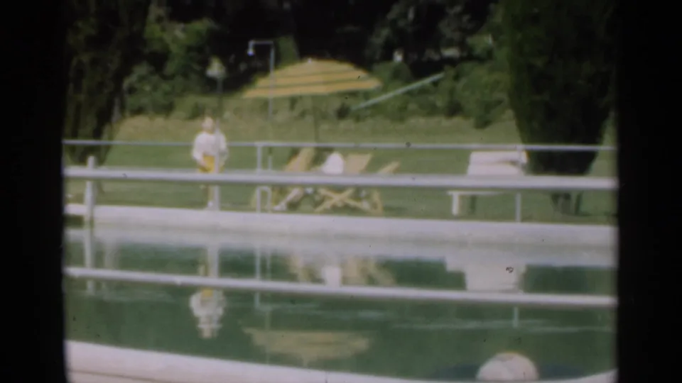 1949 Santa Cruz poolside frame with a standing figure near the water, a striped umbrella, lounge chair, lawn, and trees.
