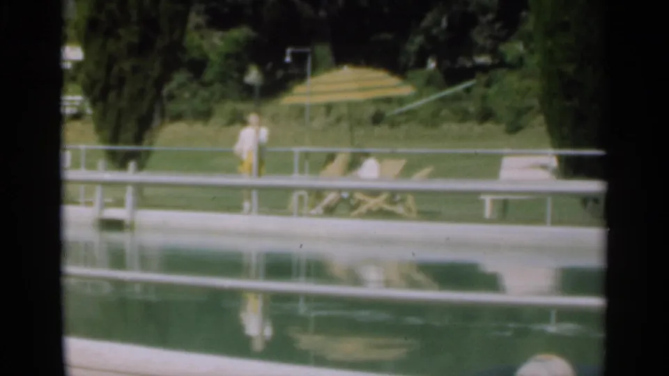 1949 Santa Cruz poolside view with a person standing near a railing and another near a lounge chair under a striped umbrella.