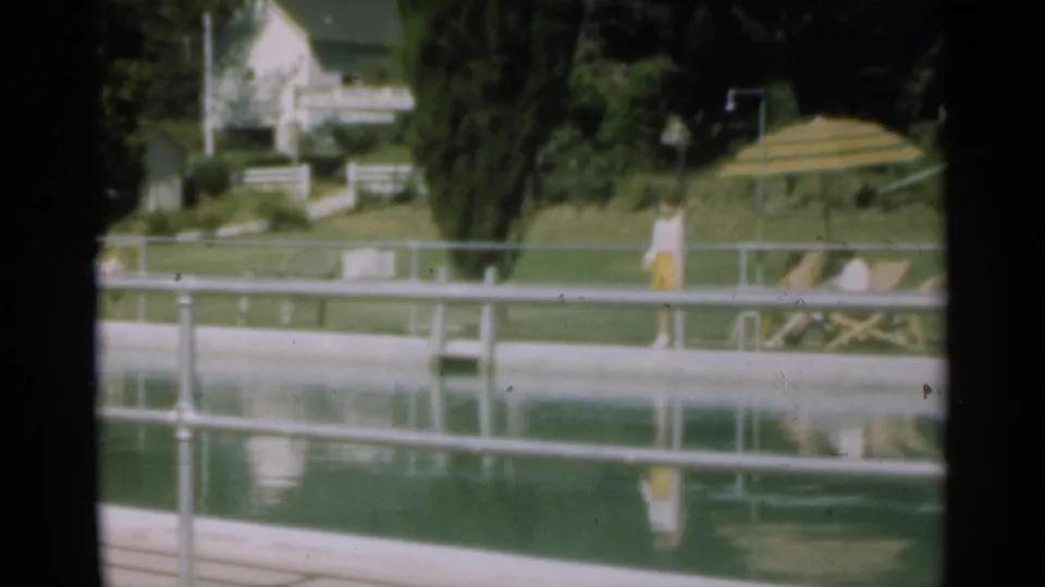 1949 Santa Cruz frame showing a poolside railing, standing figure, striped umbrella, lounge chair, trees, lawn, and distant house.