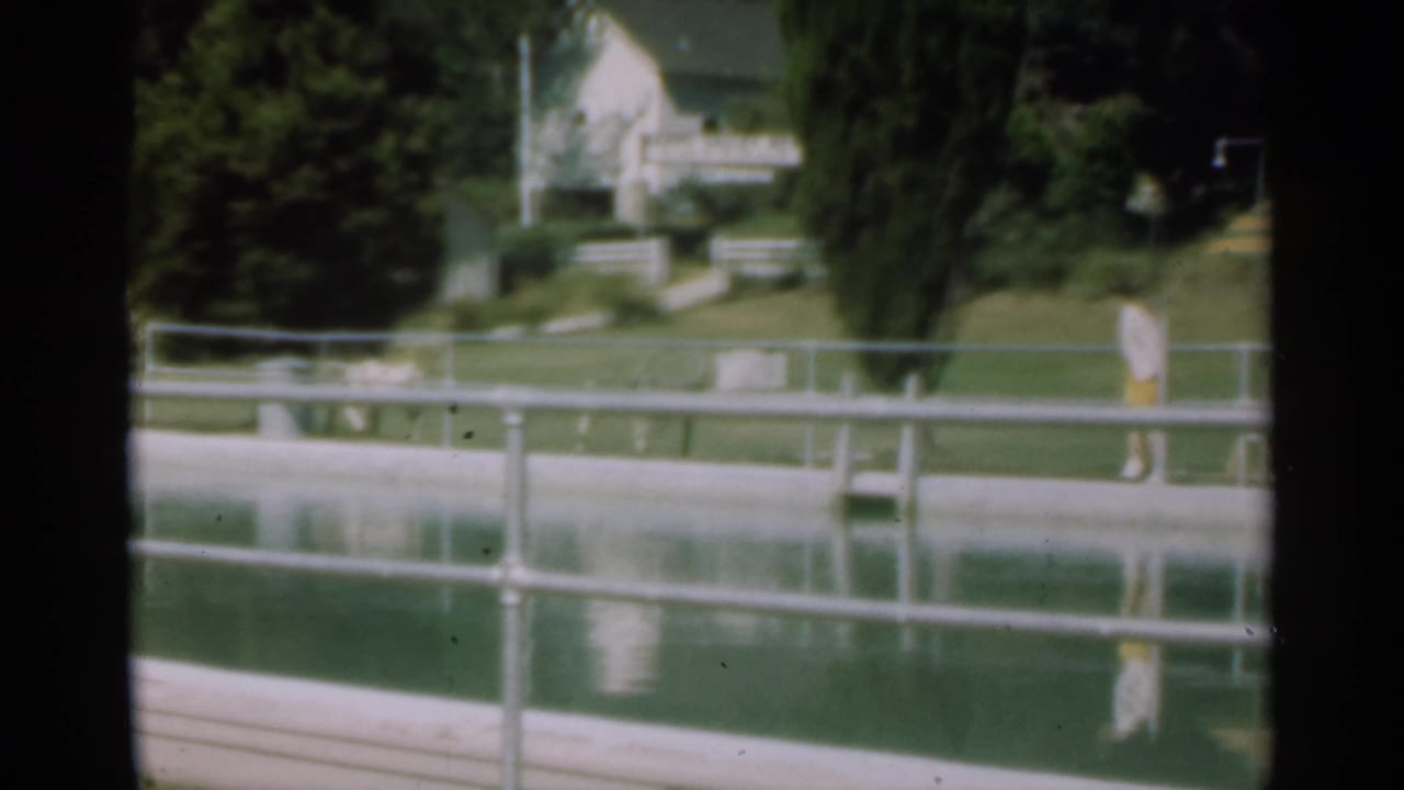 Large pond with metal railing, trees, and a white house beyond the water.