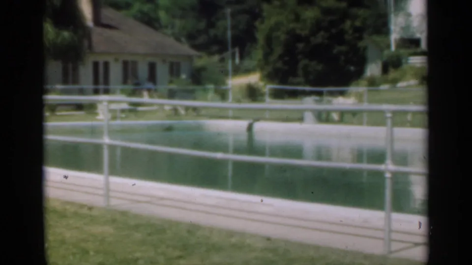 1949 Santa Cruz frame showing a house behind a railed pond, with trees and soft reflections.