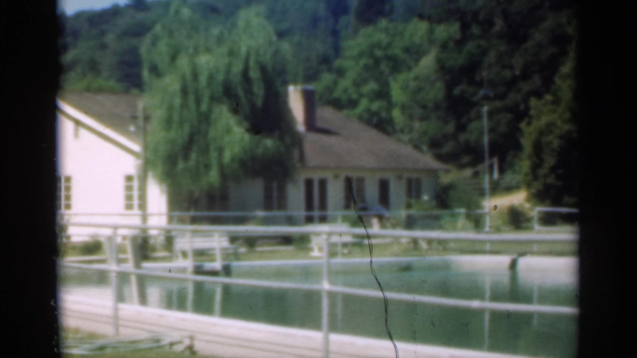 Light farmhouse beyond a pond with railings and a large willow tree.