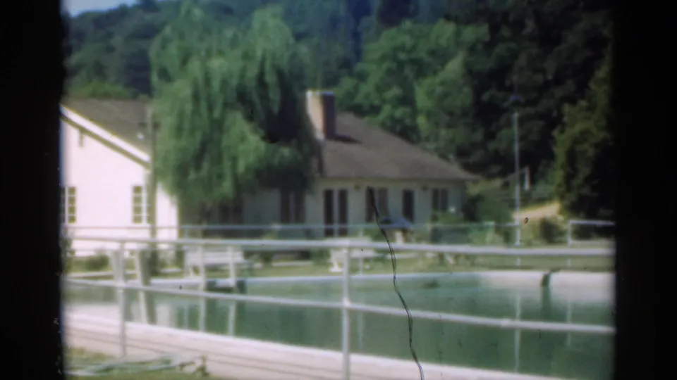 1949 Santa Cruz home-movie frame showing a light farmhouse, pond or pool, white railings, willow tree, and wooded background.