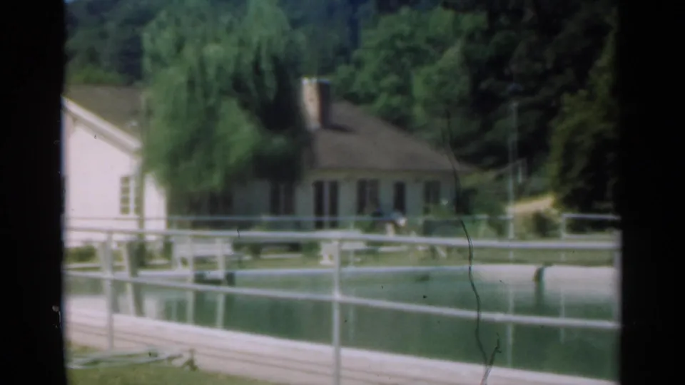 A soft 8mm frame shows a light farmhouse partly obscured by a willow, with a pond or pool and railings in the foreground.