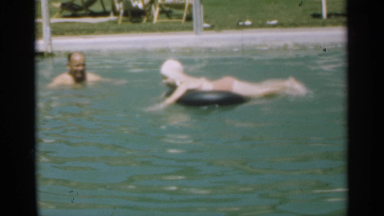 Adult swimmer watches a young swimmer on a black inner tube in an outdoor pool.