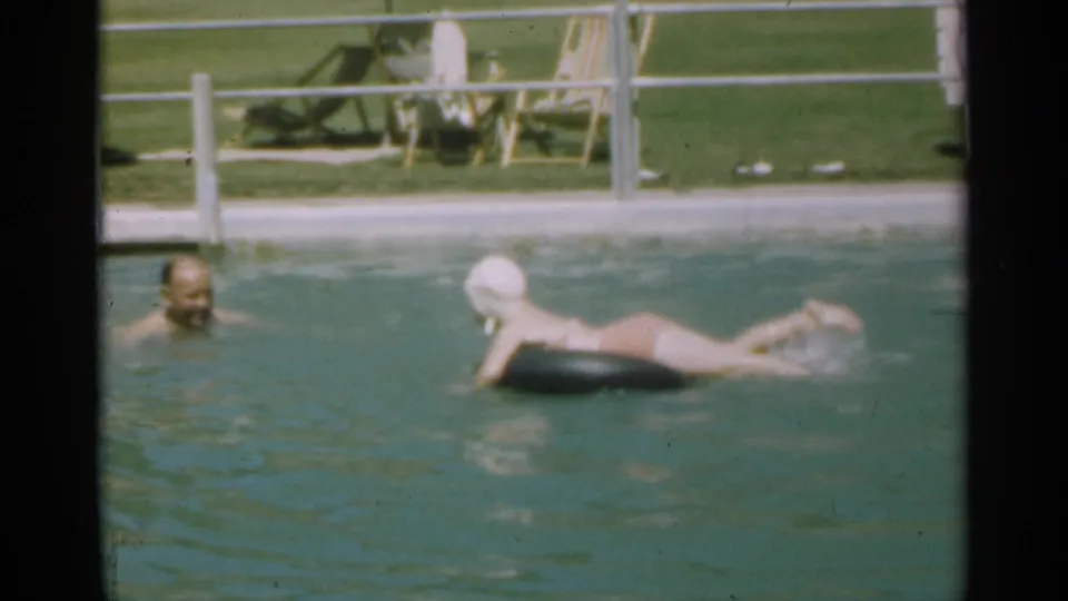 1949 Santa Cruz home-movie frame showing the adult and young swimmer in the pool, with a fence, grass, and lounge chairs behind them.