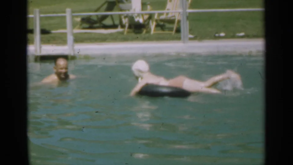 1949 Santa Cruz pool footage frame with a young swimmer on a black inner tube facing an adult, with pool fencing and lawn visible beyond.