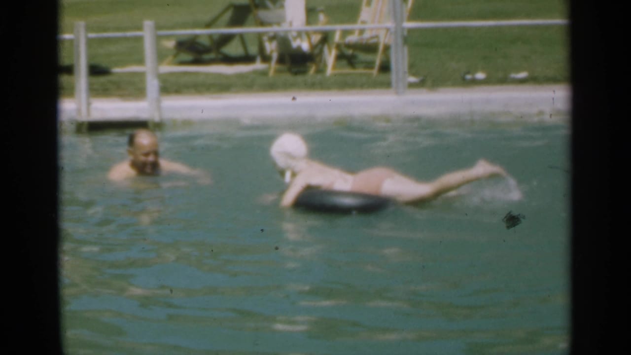 Man wading in a pool while a swimmer in a white cap floats on a black inner tube.