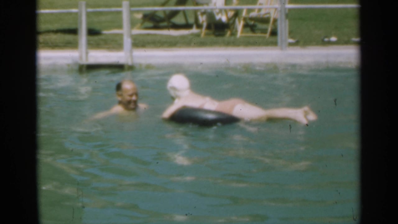 Older man swims beside a woman floating on a black inner tube in a pool.