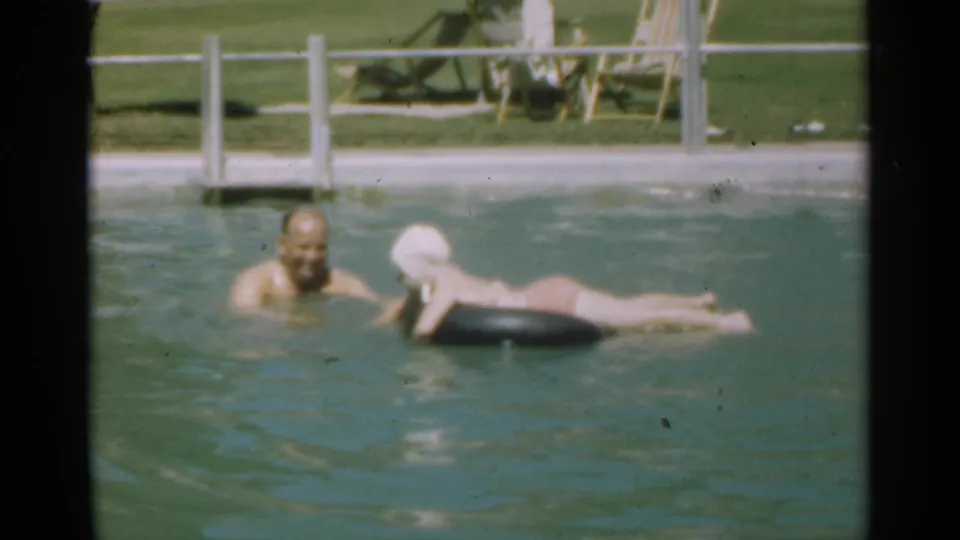 The older man faces the woman as she rests on a black ring in the Santa Cruz pool scene, with railings and lawn in back.