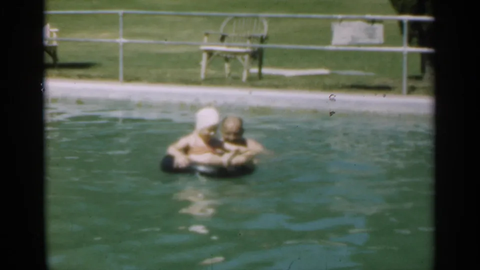 An older couple embraces around a black pool float in Santa Cruz, with railings, lawn, and lounge furniture visible beyond the water.
