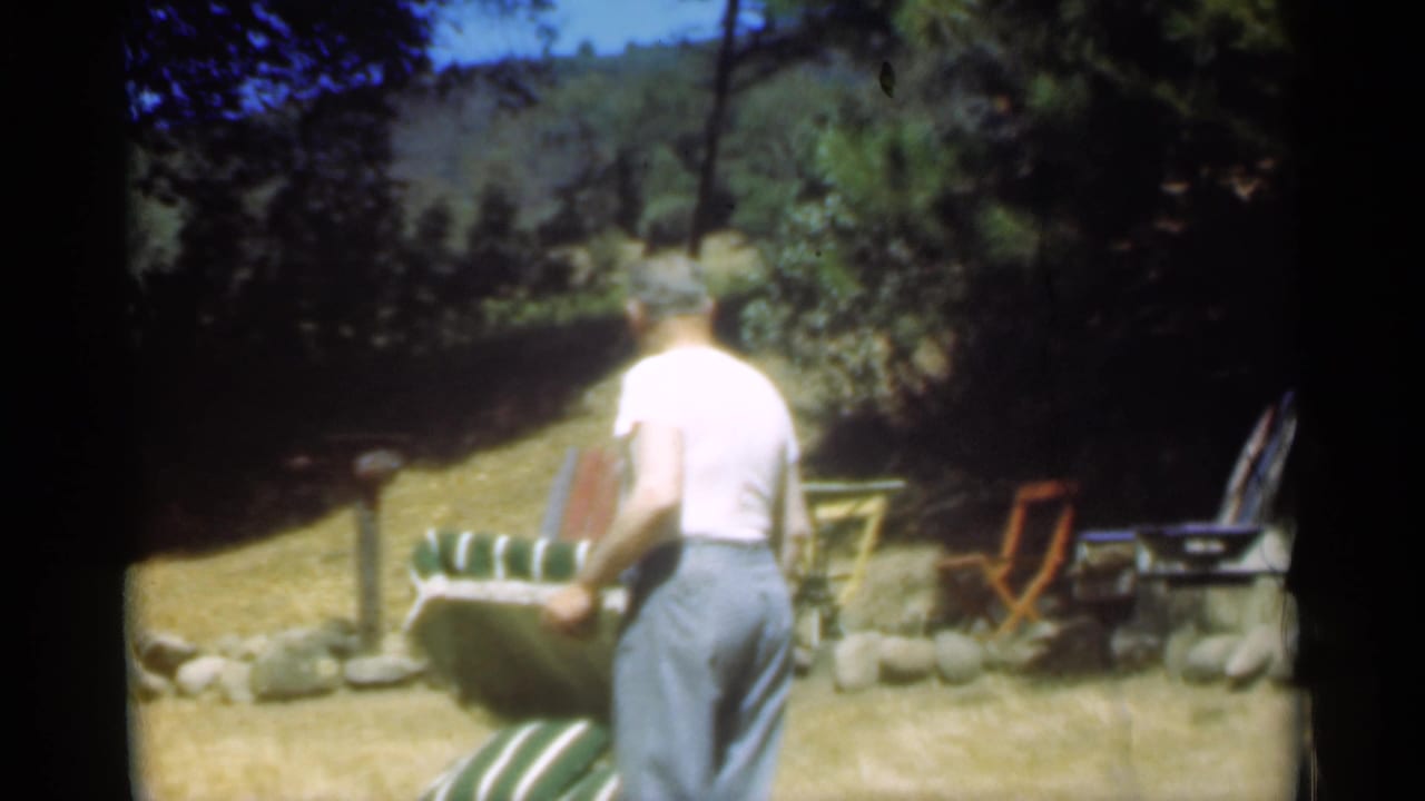 The man appears to adjust the top of a green and white striped chair in the Santa Cruz yard, with trees and outdoor furniture behind him.