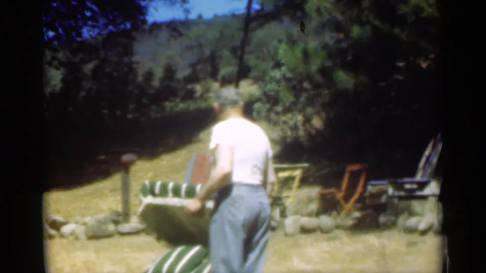 The man appears to adjust the top of a green and white striped chair in the Santa Cruz yard, with trees and outdoor furniture behind him.