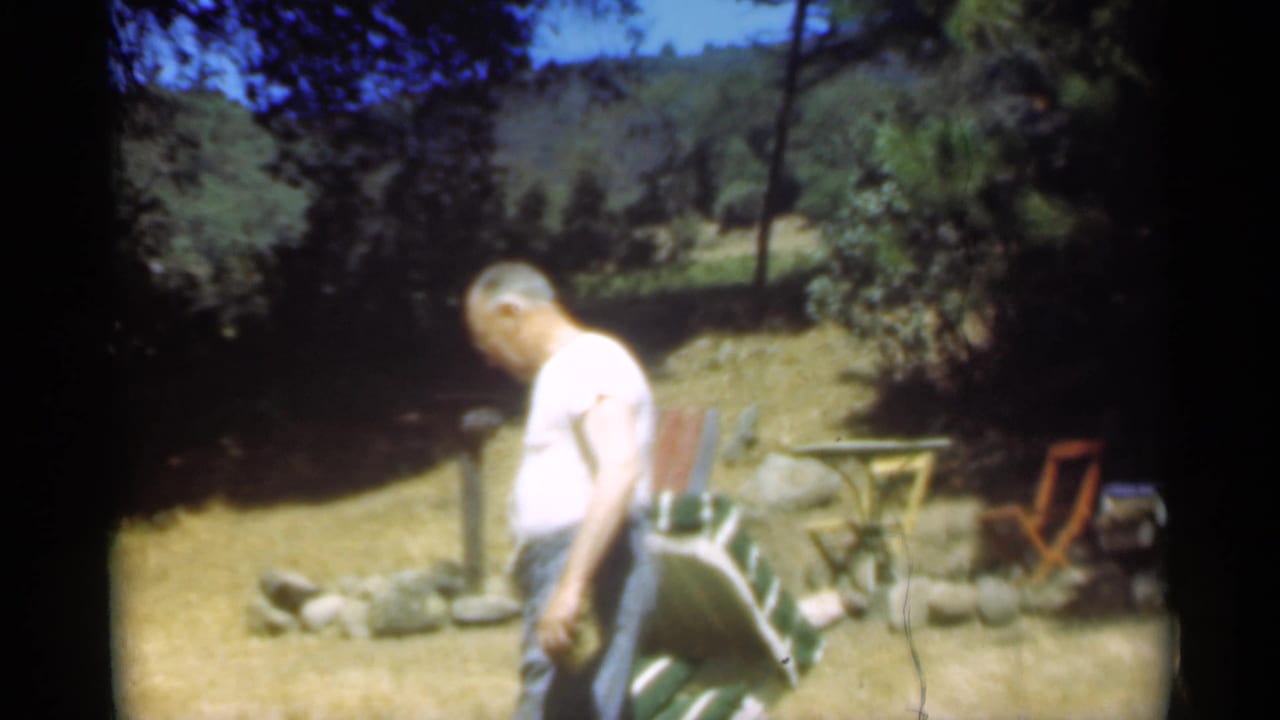 Man in a light shirt walks across a sunny lawn near a striped reclining chair.