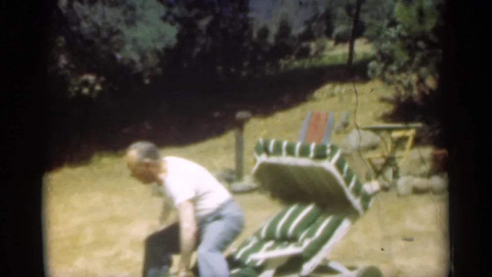 The man bends down beside a green and white striped reclining chair in a sunny Santa Cruz outdoor setting.