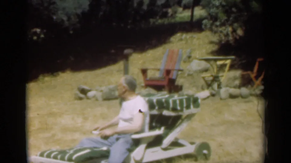 In the 1949 color 8mm scene, the man sits in the striped lawn chair with dry grass, rocks, and other chairs behind him.