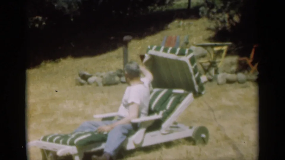 The man reclines in the striped lawn chair in Santa Cruz, with sunlit grass, trees, rocks, and outdoor furniture visible around him.