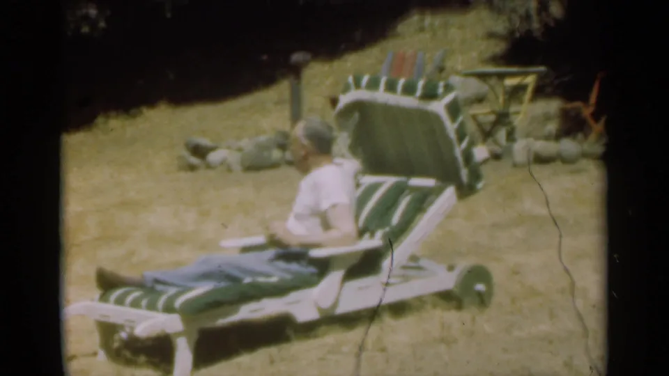 The man shifts on the striped lounge chair in a Santa Cruz yard, with a stone border and small table behind him.