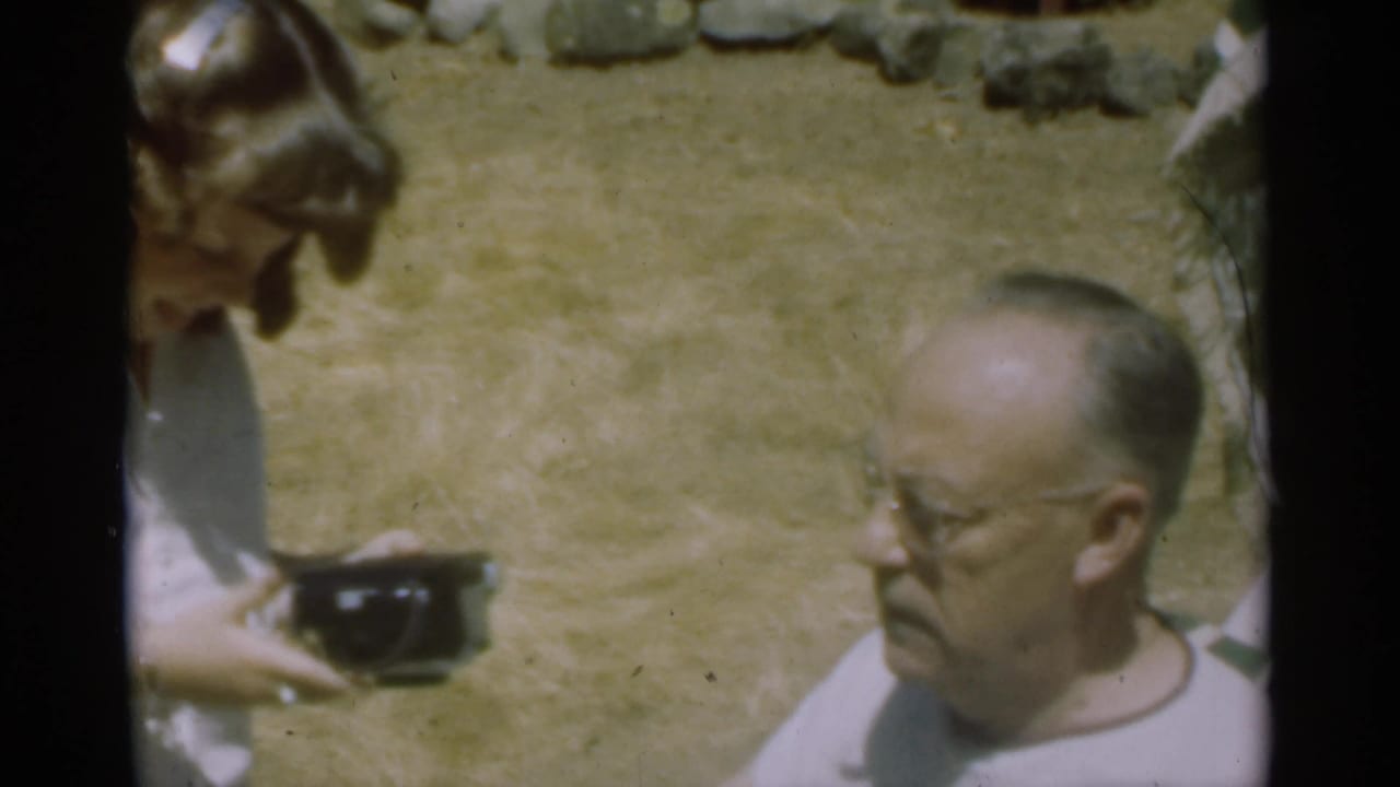 Woman holding a dark camera beside an older man with glasses on dry grass.