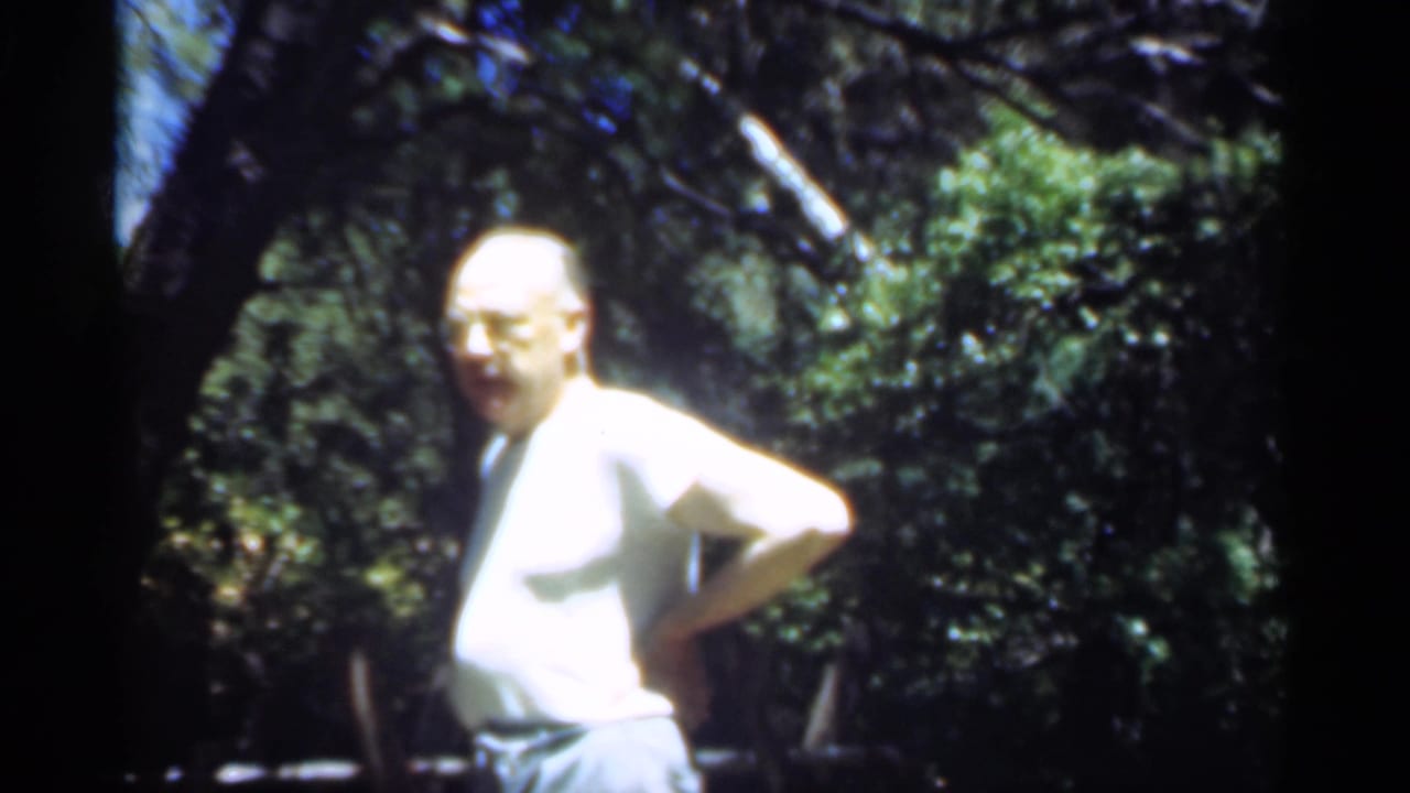 Man in a white shirt stands in wooded shade with hands on hips.