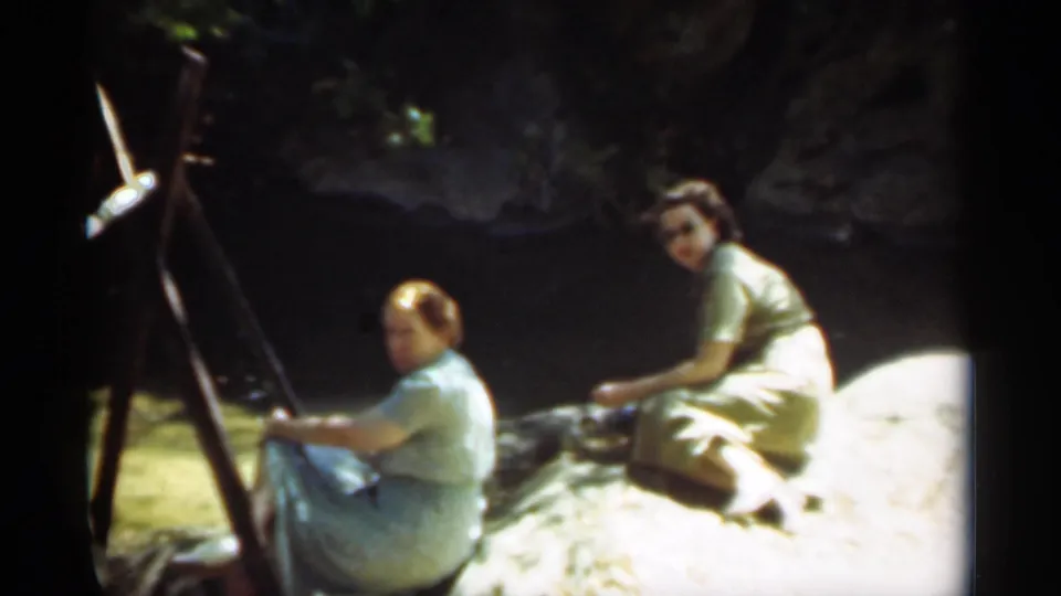 Two women in period dresses sit beside a river in the 1949 Santa Cruz home movie, with one near a wooden easel.