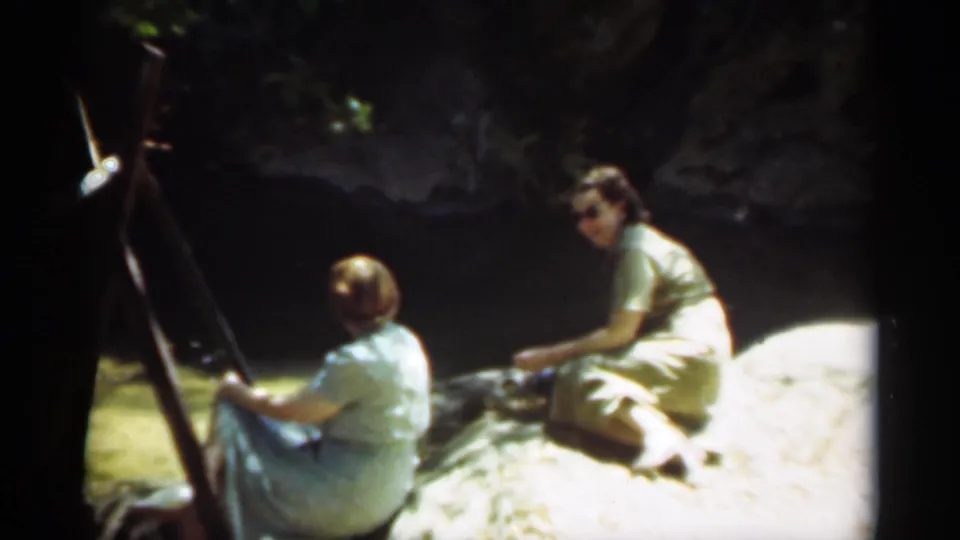 The riverside scene continues with two seated women, one looking back toward the camera, beside rocks and shaded water.