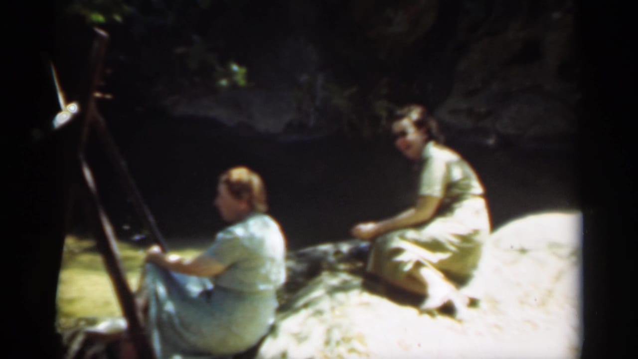 Two women sit on rocks beside shaded water, with one near an easel-like support.