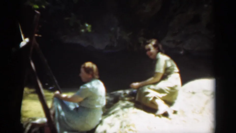 In Santa Cruz, California, two women in vintage dresses sit by shaded water on a sunny rock, one near an easel or support.