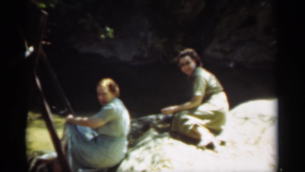 Two women seated on a rock by a shaded river in Santa Cruz appear to glance toward the camera during a quiet outdoor outing.