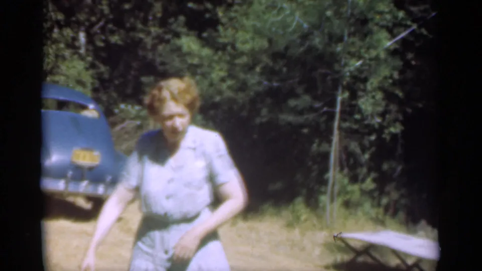 Woman beside a parked blue car in the 1949 Santa Cruz vacation footage, with trees and brush behind her.