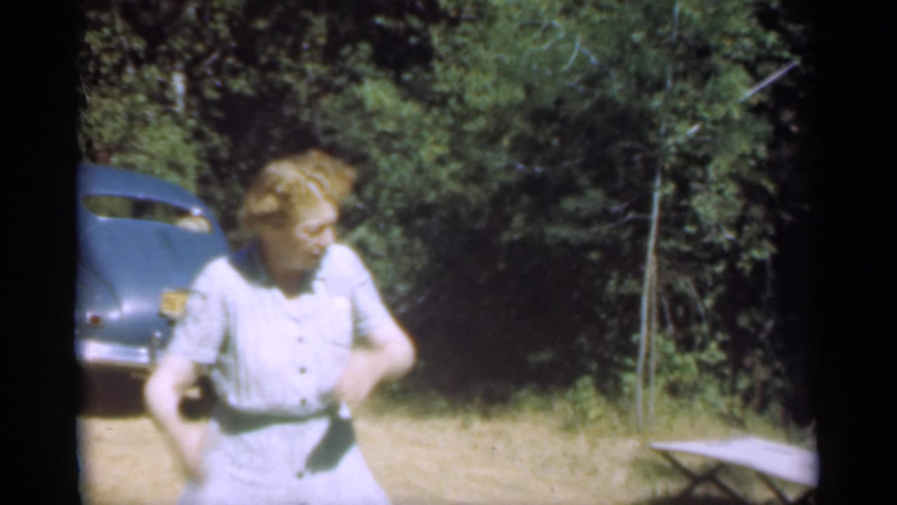 Woman in a pale dress stands near a blue vintage car beside wooded brush.