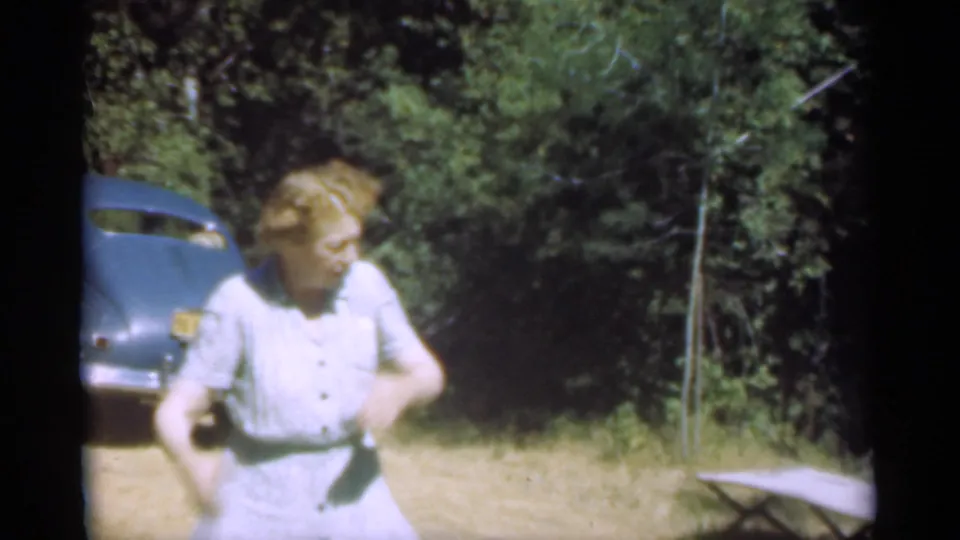 In the 1949 Santa Cruz home-movie footage, a woman in a pale dress stands outdoors near a blue vintage car and trees.