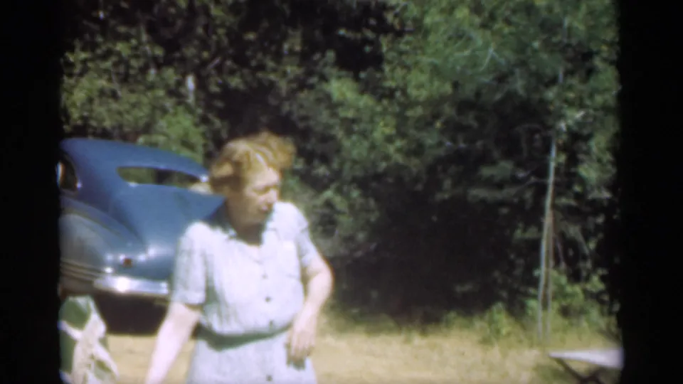 The woman appears turned to one side near the vintage blue car, with dense trees and dry ground visible in the home-movie frame.