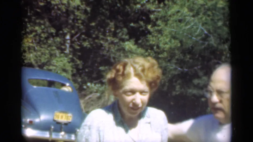 A man in a white shirt enters close beside the woman and appears to comfort her near the blue car in the 1949 Santa Cruz clip.